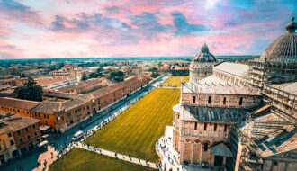 Vue panoramique aérienne de la Piazza dei Miracoli à Pise au coucher du soleil, illustrant la Cathédrale et les toits historiques de la cité sous un ciel coloré en Toscane.