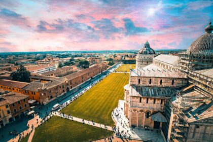 Vue panoramique aérienne de la Piazza dei Miracoli à Pise au coucher du soleil, illustrant la Cathédrale et les toits historiques de la cité sous un ciel coloré en Toscane.
