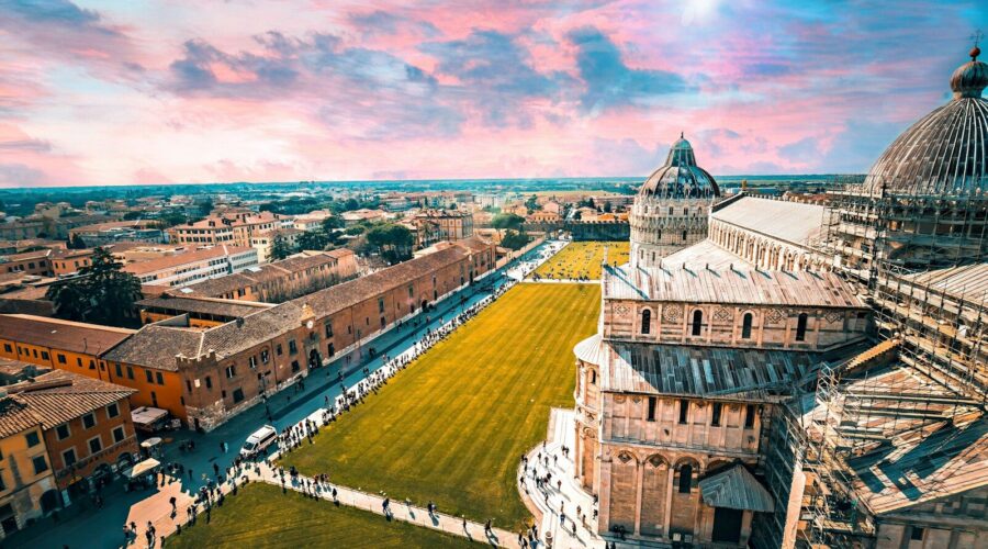 Vue panoramique aérienne de la Piazza dei Miracoli à Pise au coucher du soleil, illustrant la Cathédrale et les toits historiques de la cité sous un ciel coloré en Toscane.