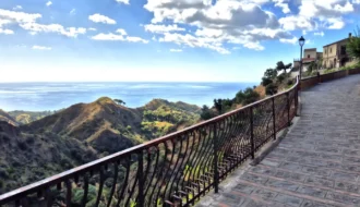 Vue panoramique depuis un chemin pavé à Savoca en Sicile, surplombant les collines verdoyantes et la mer Ionienne sous un ciel nuageux.