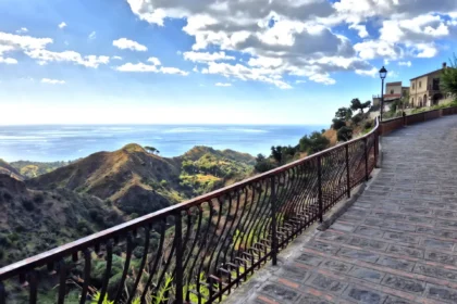 Vue panoramique depuis un chemin pavé à Savoca en Sicile, surplombant les collines verdoyantes et la mer Ionienne sous un ciel nuageux.