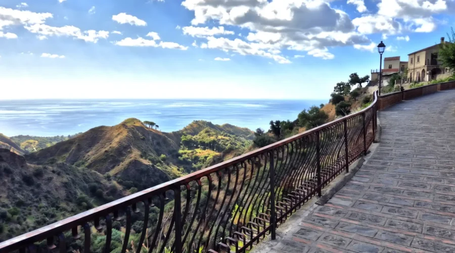 Vue panoramique depuis un chemin pavé à Savoca en Sicile, surplombant les collines verdoyantes et la mer Ionienne sous un ciel nuageux.