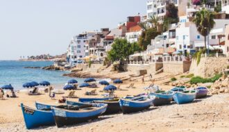 Panorama de la plage principale de Taghazout au Maroc avec ses barques de pêcheurs bleues traditionnelles sur le sable et ses maisons blanches en bord de mer.